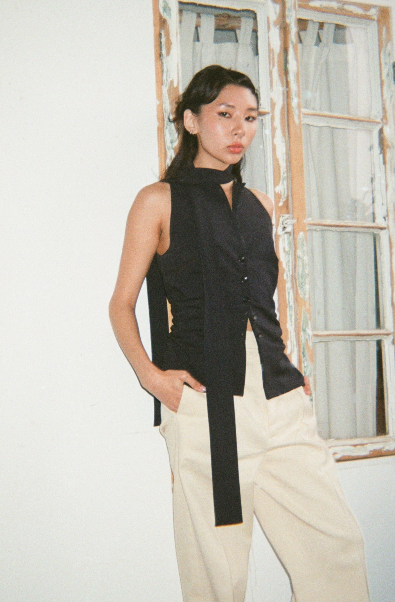 Fashionable sleeveless black shirt with a unique long necktie, worn by a model with beige pants against a white wall.