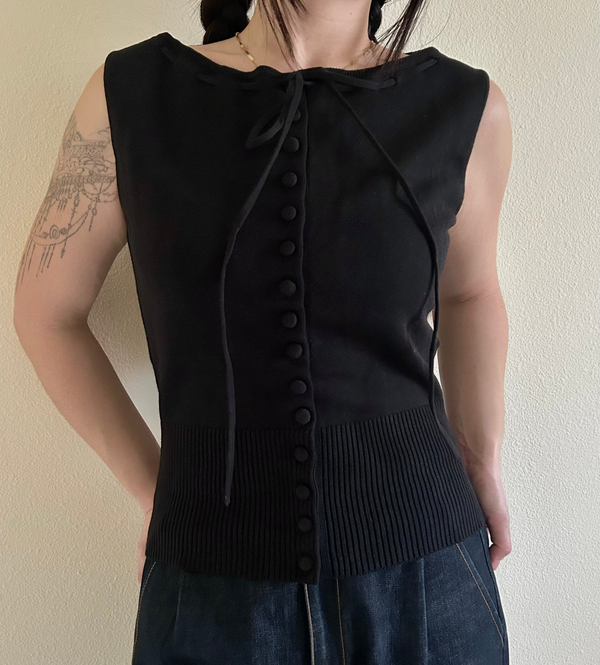 Person wearing a black sleeveless top with buttons against a plain background