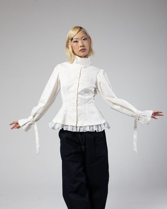 Person wearing a white blouse with ruffled details and black pants on a plain background