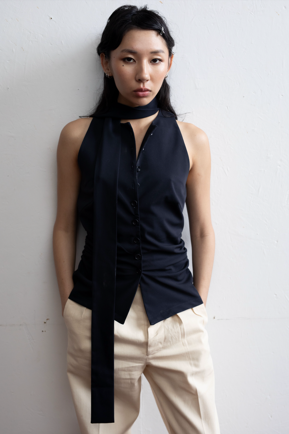 Model wearing a sleeveless black button-up shirt with a long necktie detail, paired with beige pants, posing against a white wall.