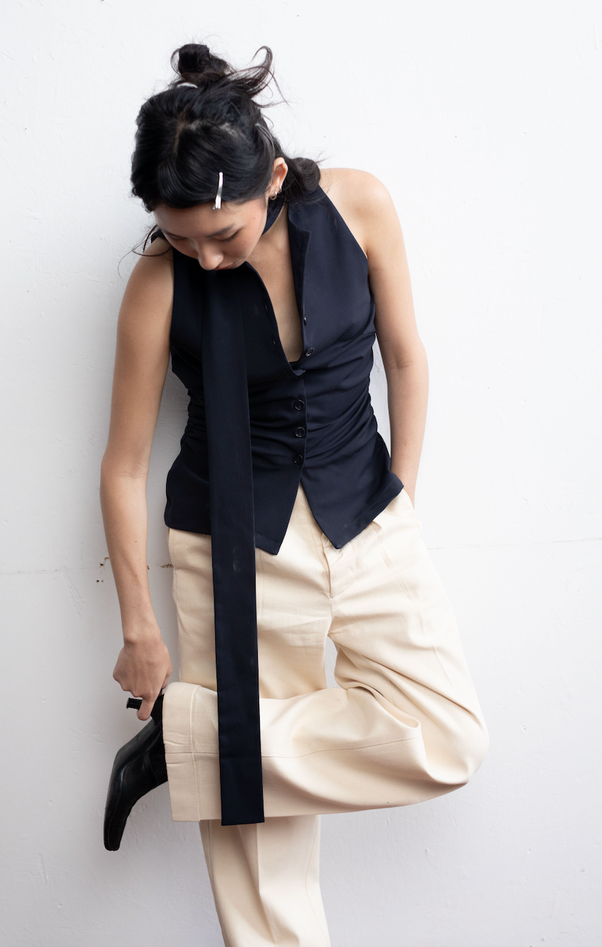 Woman in a sleeveless black button-up shirt with an extended scarf, styled with beige trousers, standing against a white backdrop.